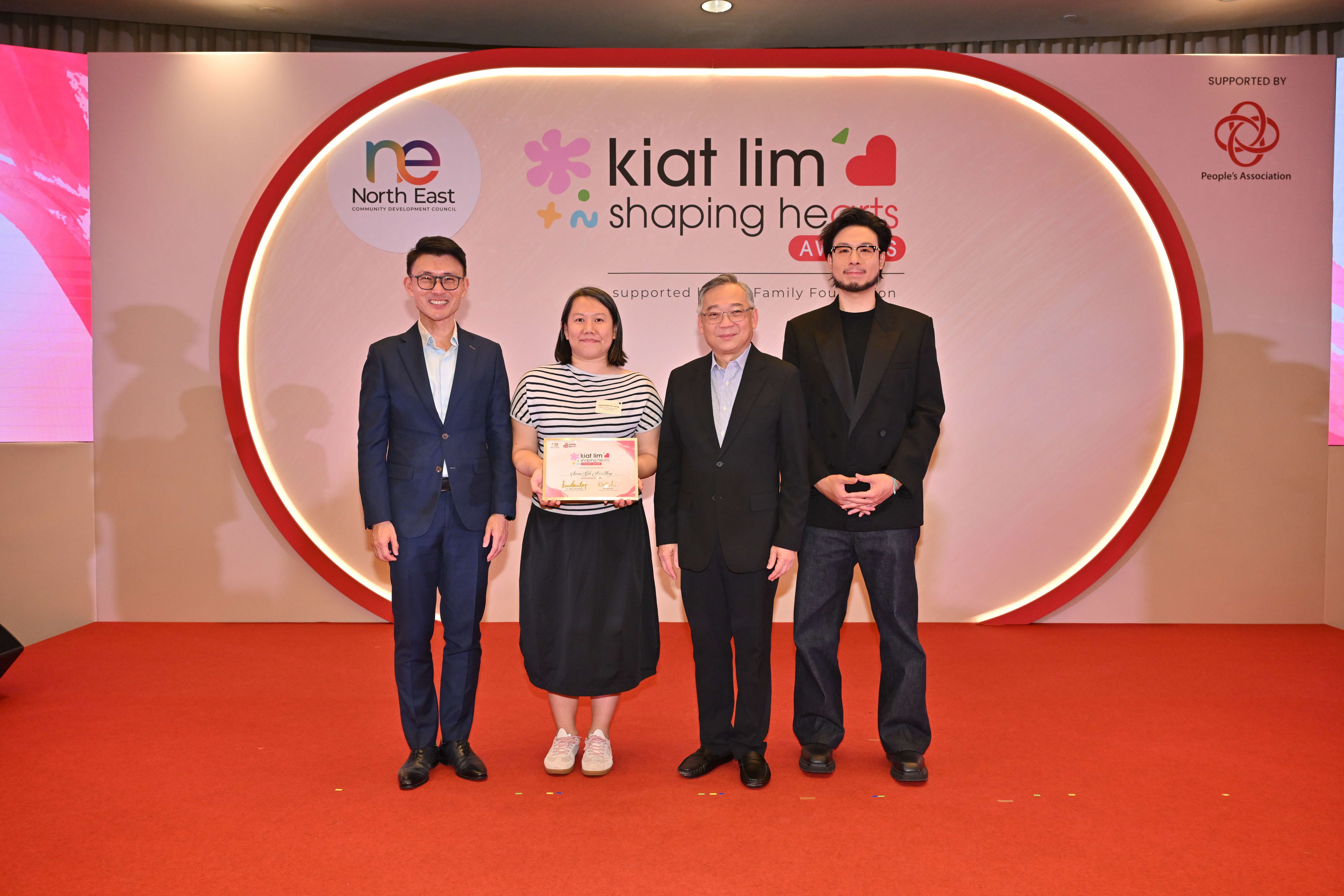 Award recipient standing on stage with the presenter, holding their certificate and posing for a group photo during the award ceremony, with a backdrop reading ‘Kiat Lim-Shaping Hearts Awards'.